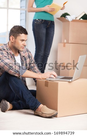 Surfing the net in a brand new house. Confident young man sitting on the floor and working on laptop while woman unpacking a carton box on the background