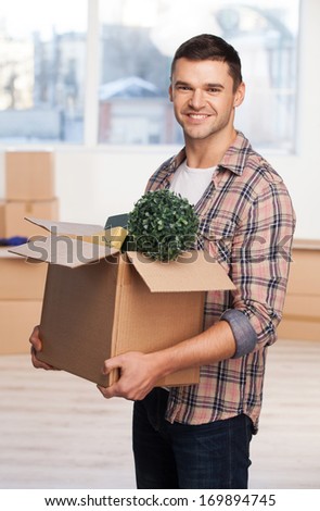 Just moved in a new house. Handsome young man holding an cardboard box with home stuff in it and smiling while more carton boxes laying on background