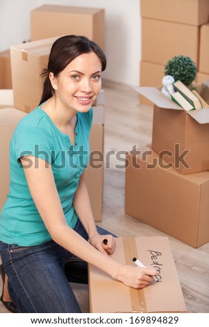 Marking a carton box. Top view of cheerful young woman marking a cardboard box and looking at camera while more boxes laying on background