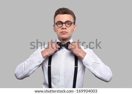 Confident in his skills. Serious young nerd man adjusting his bow tie and looking at camera while standing against grey background