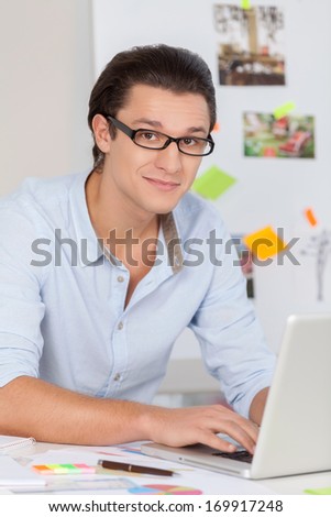 I have so many ideas. Handsome young man in glasses working on computer and smiling at camera while sitting at his working place
