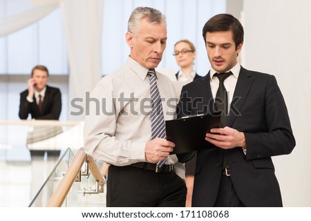 Asking an advice. Two men in formalwear discussing something while moving by staircase with people on background
