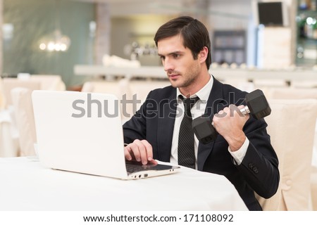 Sports and business. Confident young man in formalwear sitting at laptop and holding a dumbbell