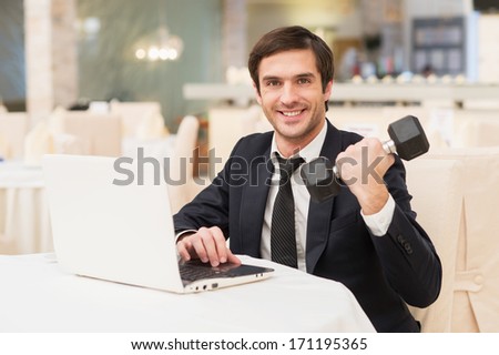Sports and business. Smiling young man in formalwear sitting at laptop and holding a dumbbell