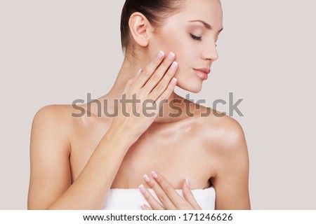Fresh and beautiful. Beautiful young woman wrapped in towel keeping eyes closed and touching her face while standing against grey background