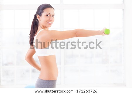 Strengthening her muscles. Side view of beautiful young Indian woman stretching out hand with dumbbell and smiling at camera