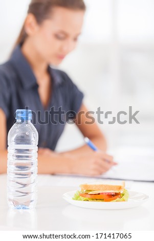 No time for lunch. Beautiful young woman writing something in her note pad while sandwich and a bottle of water laying on foreground