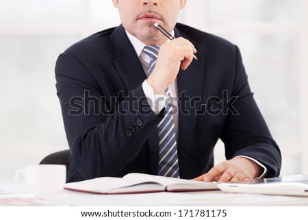 Thoughtful businessman. Cropped image of thoughtful senior man in formalwear holding pen on chin while sitting at his working place