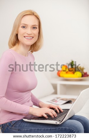 Doing business at home. Side view cheerful blond hair woman using laptop and smiling at camera
