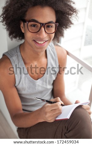 Just inspired. Top view of smiling teenage African boy sitting on the window sill and looking at camera while holding note pad in his hands