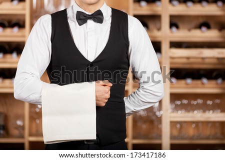 Confident waiter. Cropped image of confident young waiter standing in front of wine shelf and holding a towel on his hand