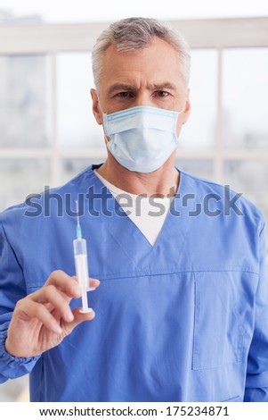 Heeling injection. Senior grey hair doctor in surgical mask holding a syringe and looking at camera