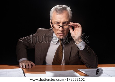 Trying to remember you. Serious senior man in formalwear sitting at his working place and adjusting his glasses while isolated on black background