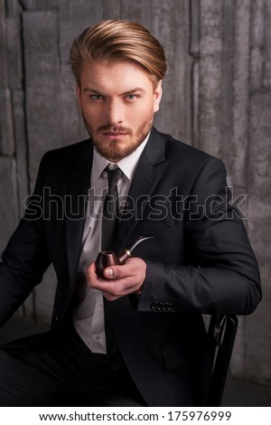 Man with a smoking pipe. Handsome young man in formalwear holding a smoking pipe and looking at camera while sitting on the chair