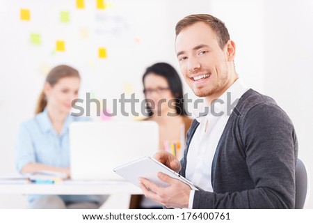 Creative team. Cheerful young man in glasses using digital tablet and smiling while his colleagues working on the background