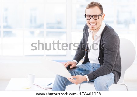 Creative executive. Handsome young man in glasses working on computer and smiling at camera while sitting at his working place