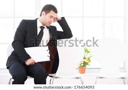 Tired of waiting. Bored young man in formalwear sitting at the chair and holding head in hand while other chairs standing in a row