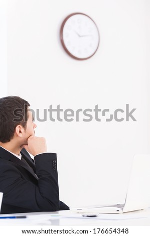 Time is gone. Worried young man in formalwear looking at the wall clock and holding hand on chin while sitting at his working place