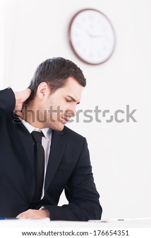 No time for break. Depressed young man in formalwear looking away and touching his neck while sitting at his working place with wall clock on background