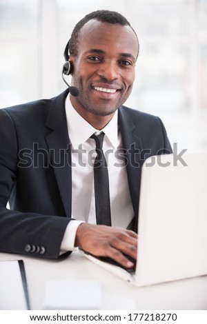 Leader of support team. Handsome young African man in formalwear and headset working on laptop and smiling at camera