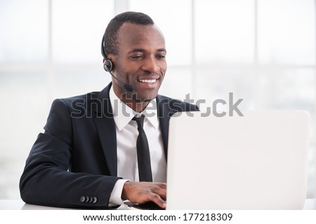 Customer service representative at work. Cheerful young African man in formalwear and headset working on laptop and smiling