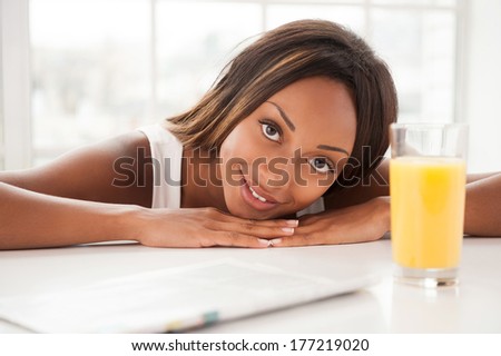 Starting day with orange juice. Beautiful young African woman looking at camera and smiling while sitting at the table with glass of orange juice on it