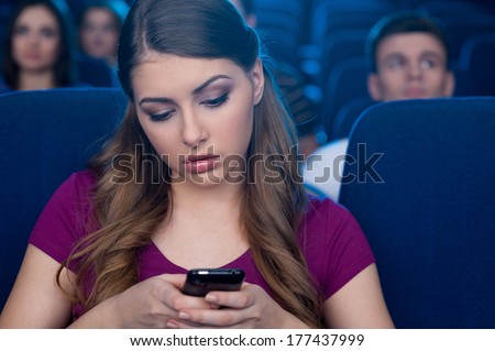 Feeling bored at the cinema. Bored young woman holding a mobile phone and looking at it while watching movie at the cinema