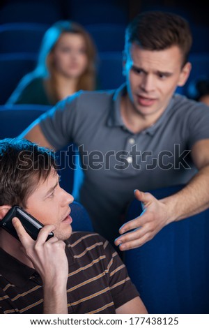 Can you stop talking? Young man asking to stop the other man talking on the mobile phone while watching movie at the cinema