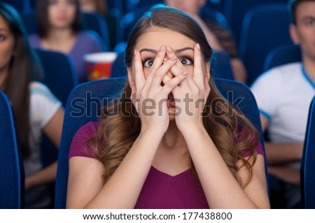 Watching a scary movie. Shocked young woman covering face with hands and watching movie while sitting at the cinema