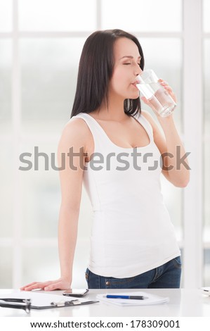 Refreshing with glass of water. Beautiful young woman drinking water while leaning at the table