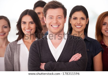 He is a real team leader. Confident young man keeping arms crossed and smiling while group of people standing on background