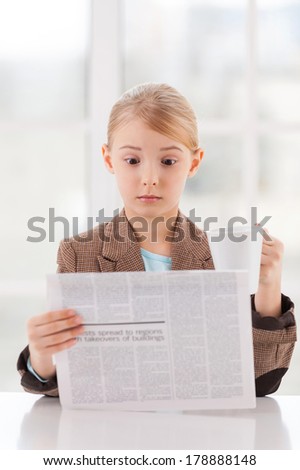 Shocking news. Surprised little girl in formalwear reading newspaper and holding a cup while sitting at the table