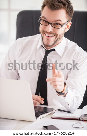 Making your business easier. Handsome young man in shirt and tie pointing you and smiling while sitting at his working place
