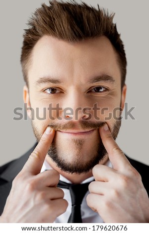 Making a fake smile. Portrait of young man in formalwear making a smile by his fingers while standing against grey background