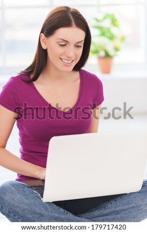 Surfing the net at home. Attractive young woman working on laptop and smiling while sitting on the floor at her apartment