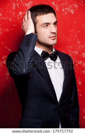 Handsome and stylish. Handsome young man in suit and bow tie touching his hair and looking away while standing against red background