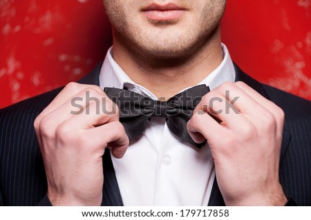 Tying his bow tie. Cropped image of handsome young man in formalwear adjusting his bow tie while standing against red background