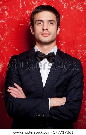 Handsome in bow tie. Handsome young man in suit and bow tie keeping arms crossed and looking at camera while standing against red background