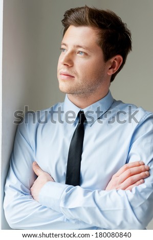 Thinking about solutions. Handsome young man in shirt and tie looking away and keeping arms crossed while leaning at the wall