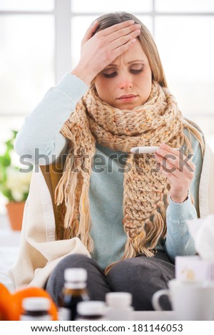 Checking body temperature. Young woman covered with plaid checking her body temperature while sitting in bed at her apartment