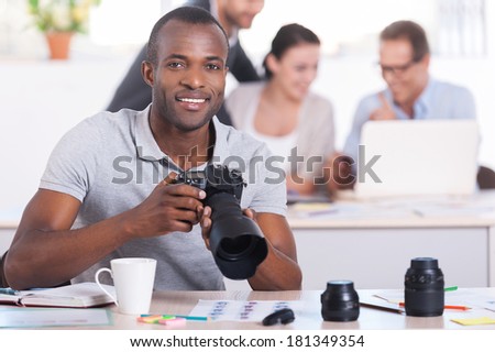 Creative people at work. Handsome young African man holding camera and smiling while three people working on background