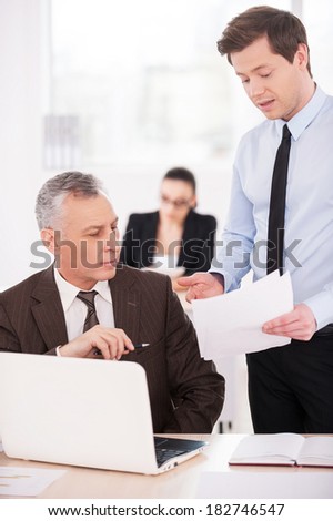 I need an advice. Two confident business people discussing something while on of them pointing paper with woman working on background