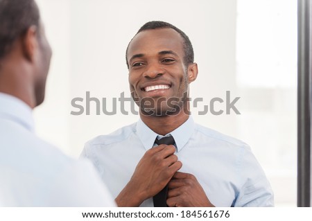 Confident about his look. Young African man adjusting his necktie while standing against mirror