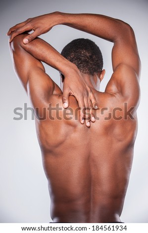 Joint pain. Rear view of young shirtless African man touching his neck and elbow while standing against grey background