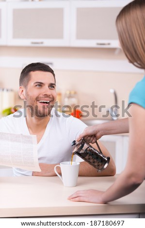 Happy Sunday morning. Woman pouring coffee to the cup while happy young man holding newspaper and smiling