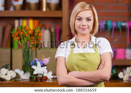 Beautiful florist at working place. Beautiful young female florist keeping arms crossed and looking at camera while standing near her working place