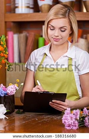 Beautiful florist at work. Beautiful young female florist writing something at note pad and taking on the phone while standing at her working place
