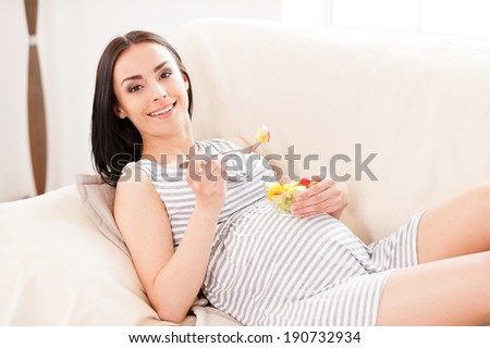 Healthy eating for both of us. Beautiful pregnant woman eating a fruit salad and smiling while lying on a couch