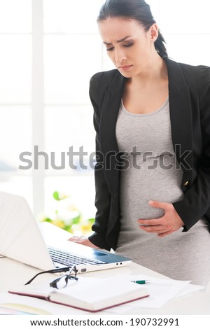 Pregnancy pains and aches. Depressed pregnant businesswoman touching her abdomen while standing near her working place in office