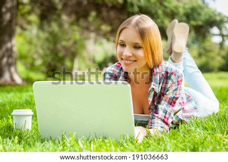 Surfing the net outdoors. Beautiful young female student working on laptop and smiling while lying on the grass in park